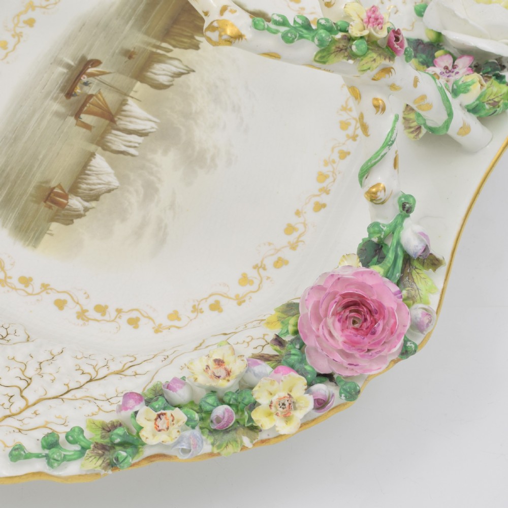 A white porcelain basket decorated with hand-sculpted flowers along the rim, a gilded twig-shaped handle, and a hand-painted marine landscape scene in the centre. View VI