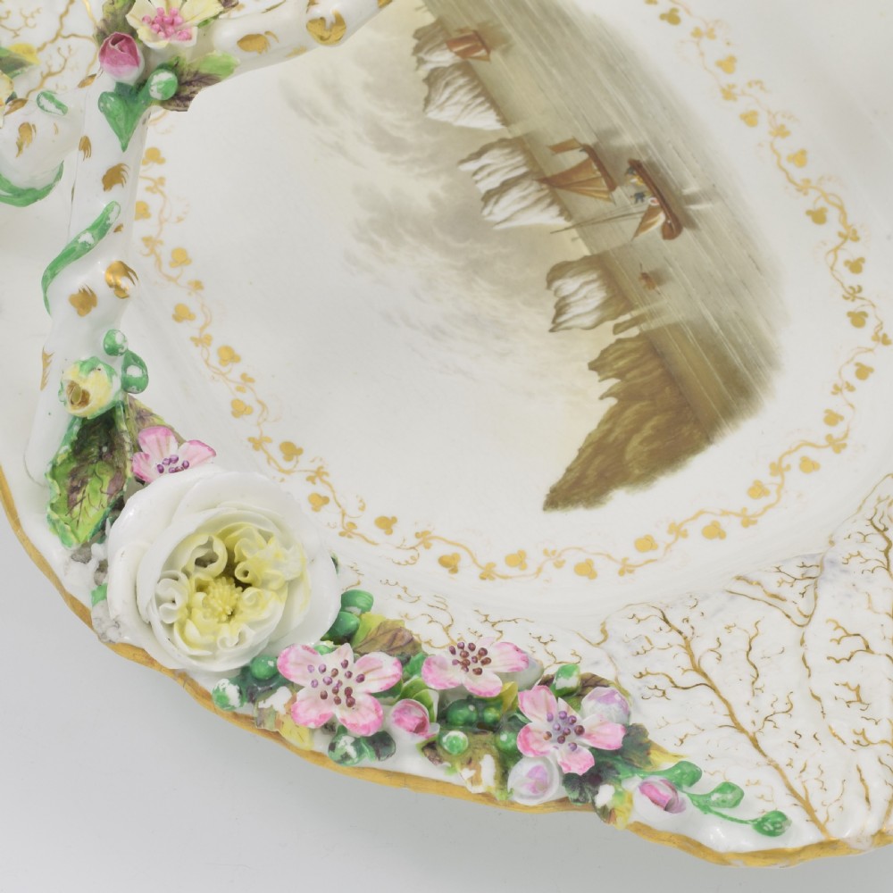 A white porcelain basket decorated with hand-sculpted flowers along the rim, a gilded twig-shaped handle, and a hand-painted marine landscape scene in the centre. View VII