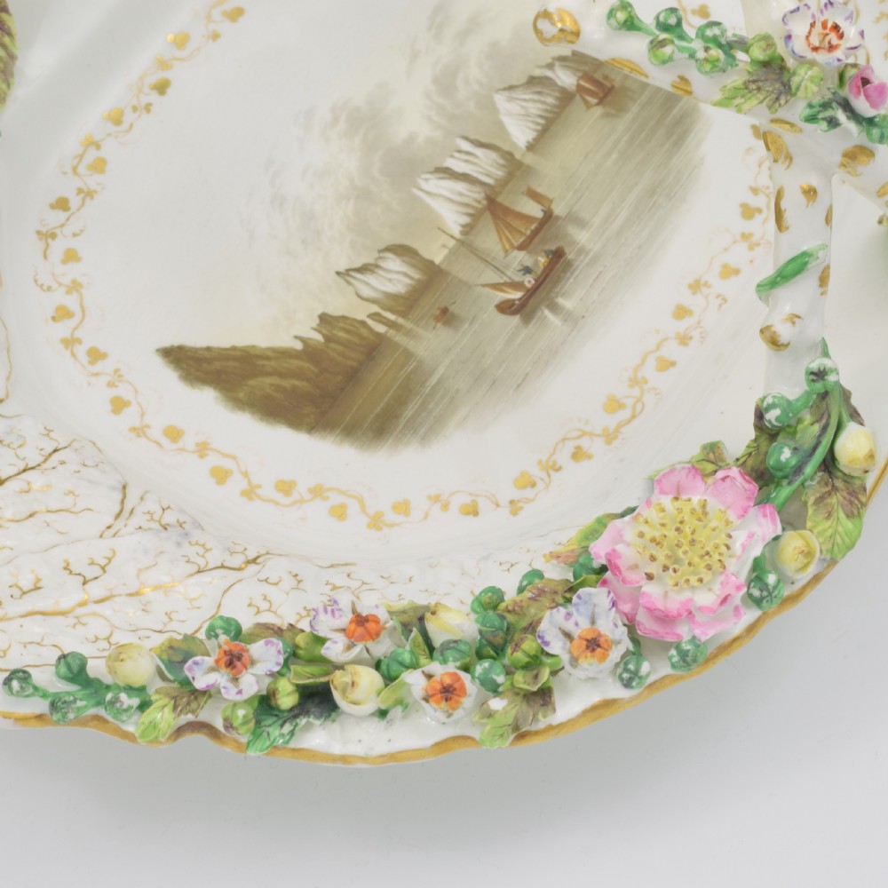 A white porcelain basket decorated with hand-sculpted flowers along the rim, a gilded twig-shaped handle, and a hand-painted marine landscape scene in the centre. View VIII