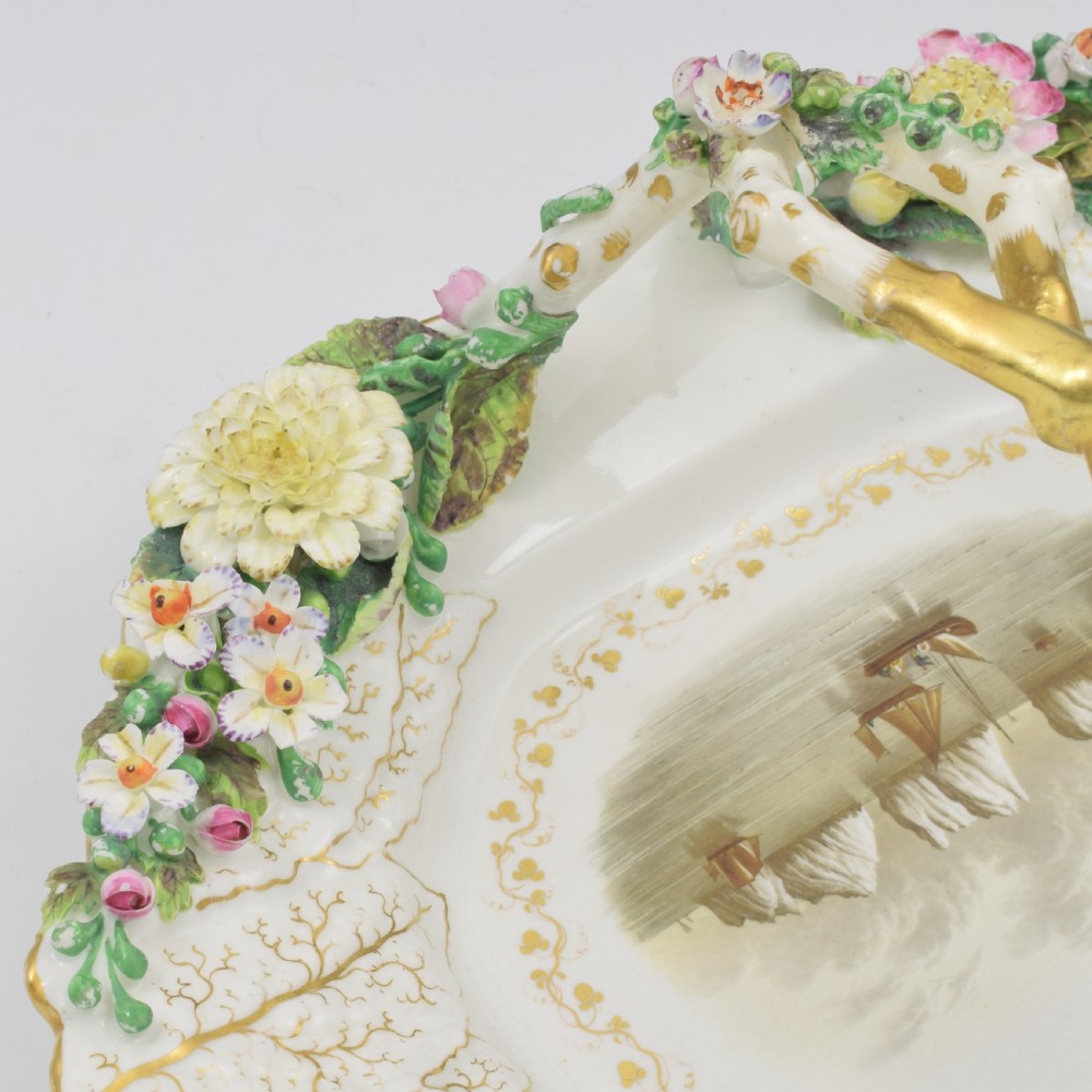 A white porcelain basket decorated with hand-sculpted flowers along the rim, a gilded twig-shaped handle, and a hand-painted marine landscape scene in the centre. View IX