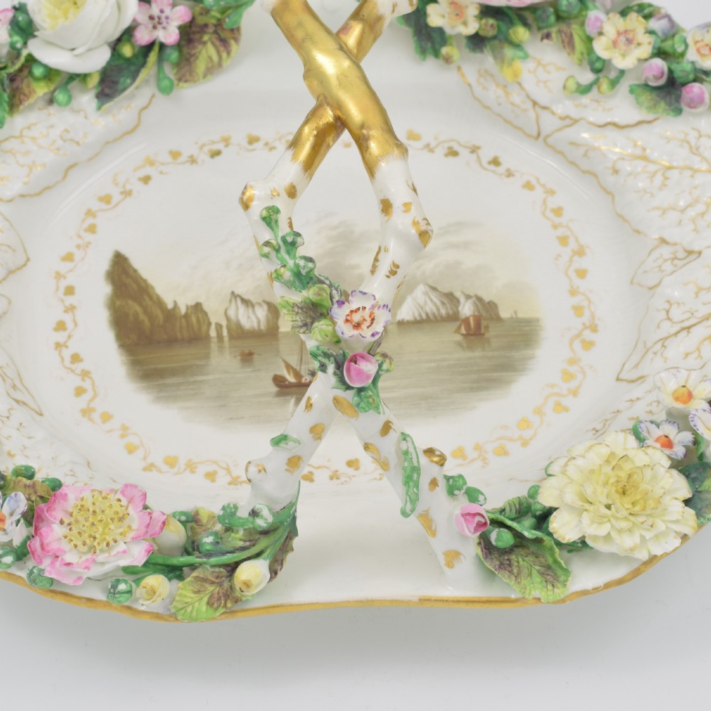 A white porcelain basket decorated with hand-sculpted flowers along the rim, a gilded twig-shaped handle, and a hand-painted marine landscape scene in the centre. View X
