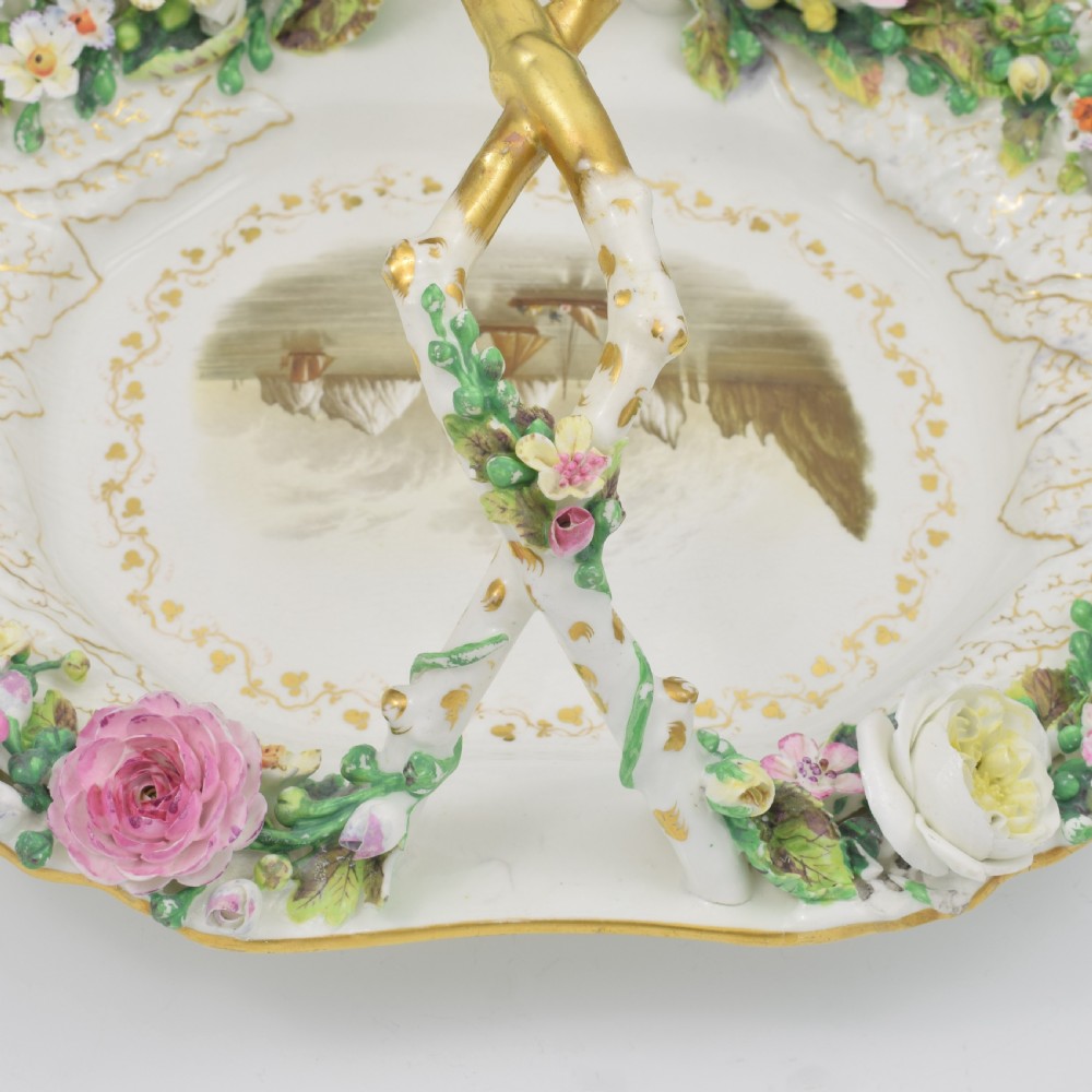 A white porcelain basket decorated with hand-sculpted flowers along the rim, a gilded twig-shaped handle, and a hand-painted marine landscape scene in the centre. View XII