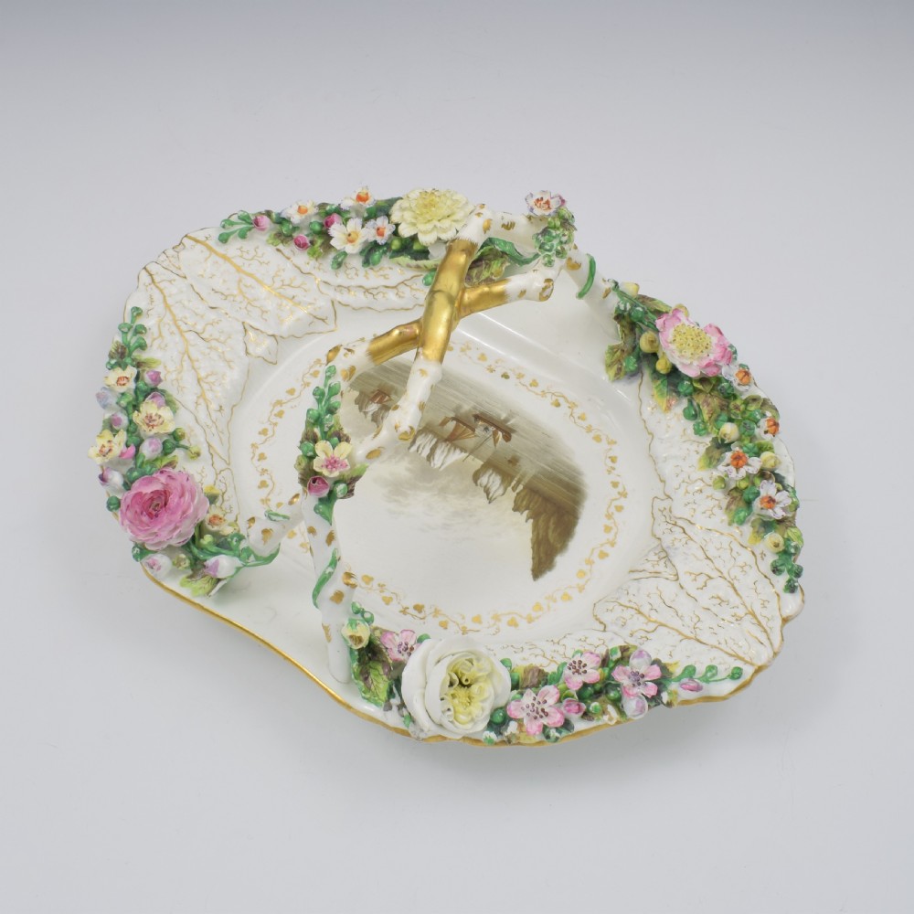 A white porcelain basket decorated with hand-sculpted flowers along the rim, a gilded twig-shaped handle, and a hand-painted marine landscape scene in the centre. View XIV