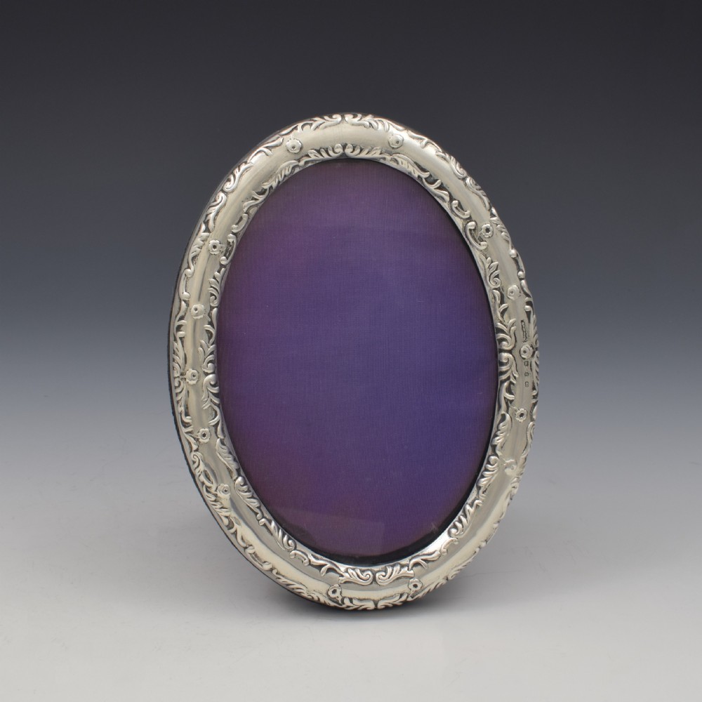 An oval silver photo frame with embossed foliate scrolling decoration surrounding a dark purple aperture.