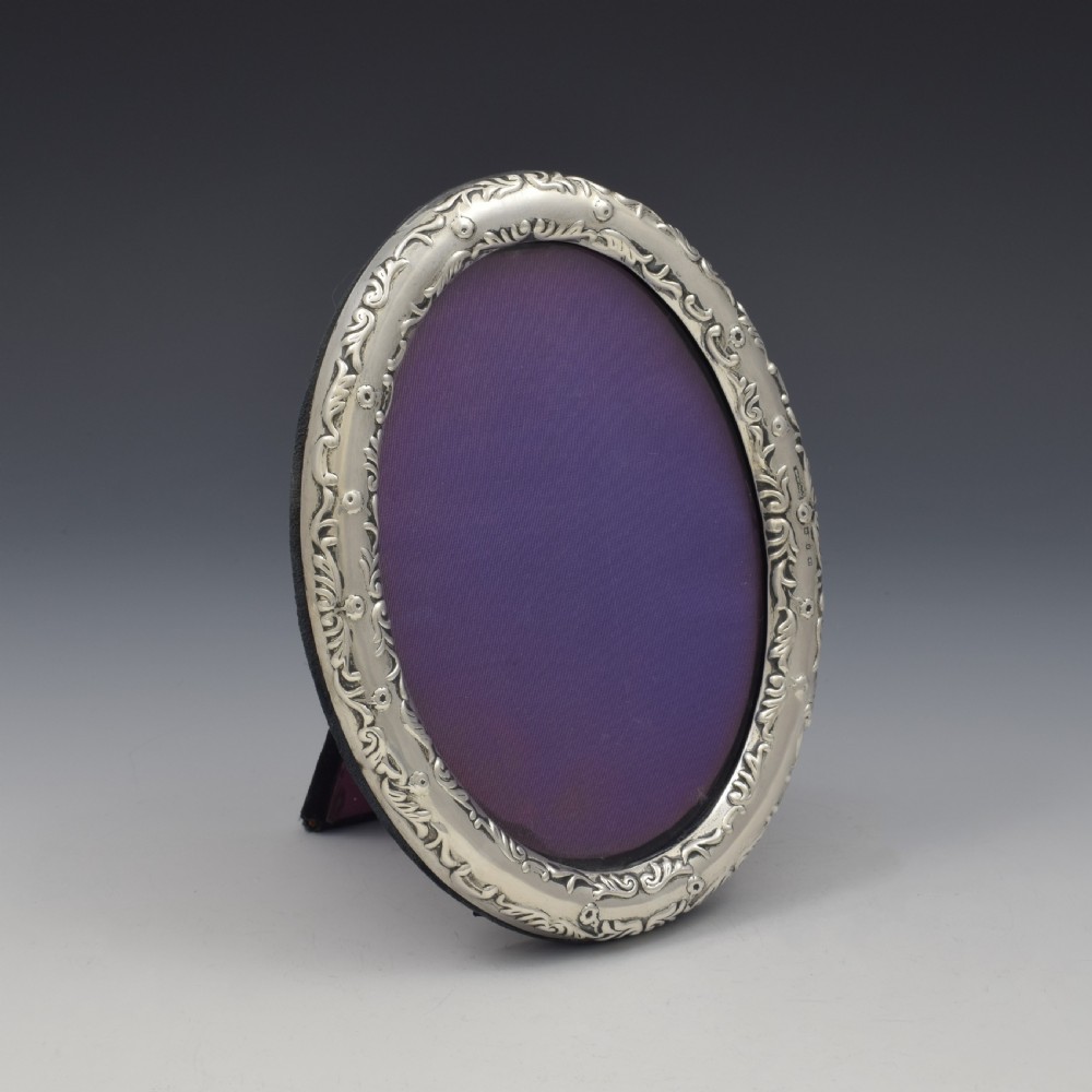 An oval silver photo frame with embossed foliate scrolling decoration surrounding a dark purple aperture. View II