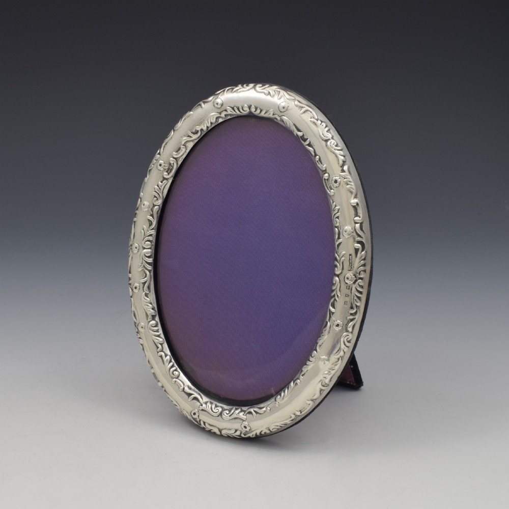 An oval silver photo frame with embossed foliate scrolling decoration surrounding a dark purple aperture. View III