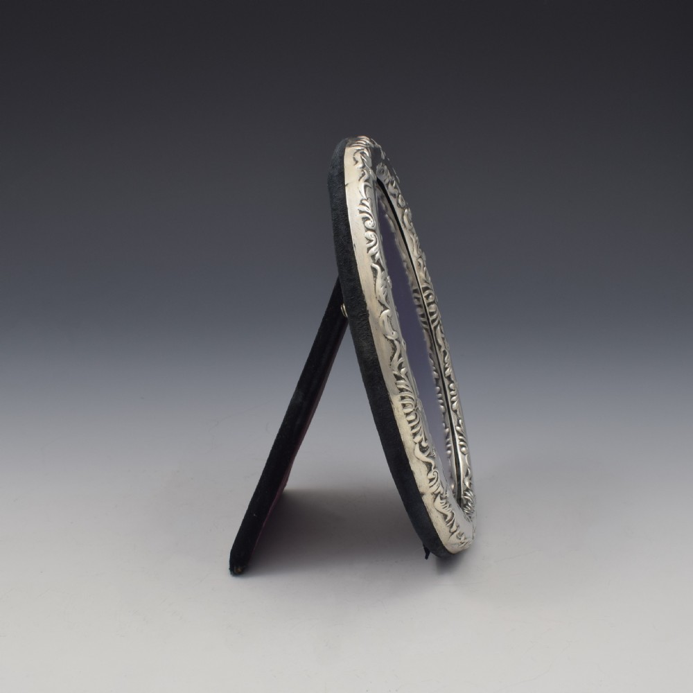 An oval silver photo frame with embossed foliate scrolling decoration surrounding a dark purple aperture. View VII