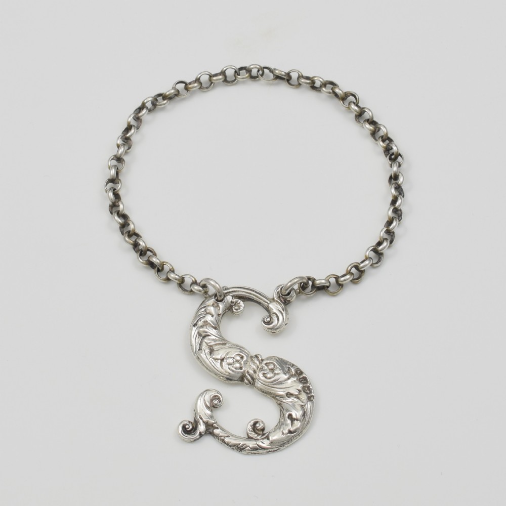 An antique Georgian sterling silver decanter label shaped as the letter S with chased foliate scrollwork and an attached chain.