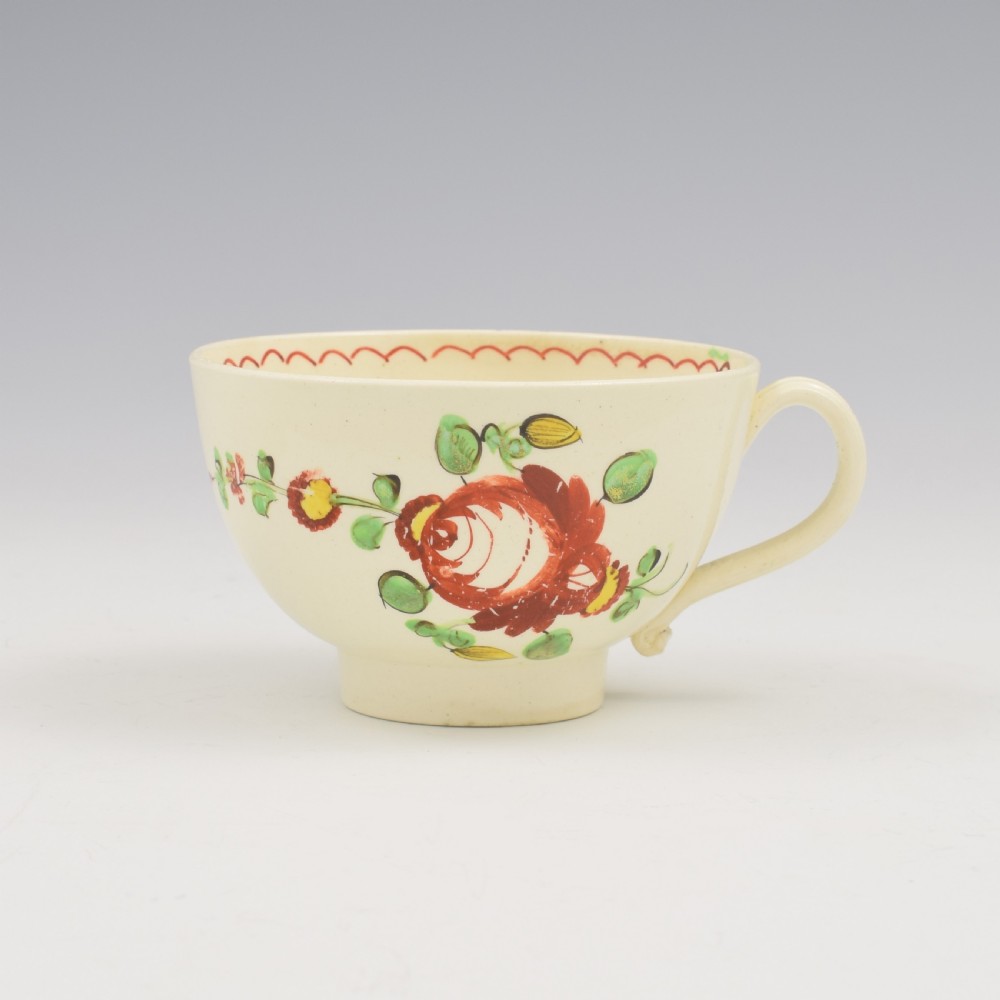 An antique creamware tea cup and saucer featuring hand-painted red, green, and yellow floral motifs in the Kings Rose pattern against a light cream backdrop. View III
