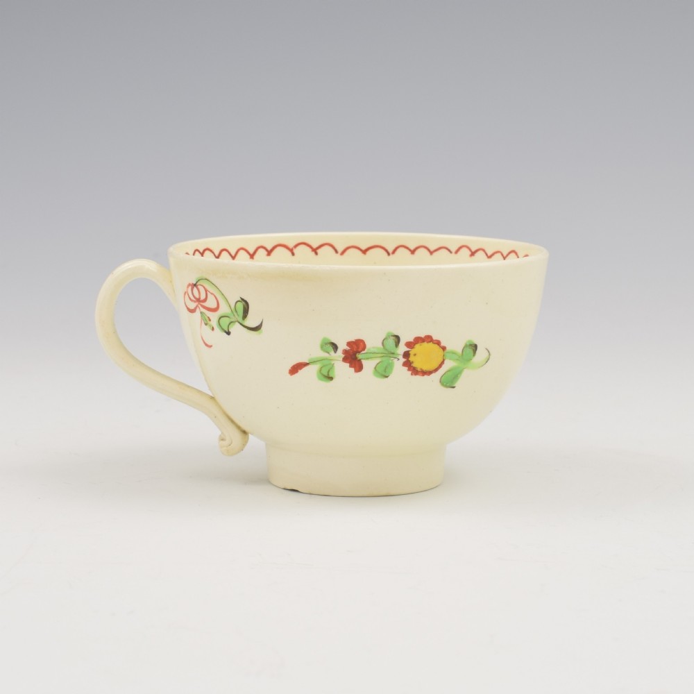 An antique creamware tea cup and saucer featuring hand-painted red, green, and yellow floral motifs in the Kings Rose pattern against a light cream backdrop. View V