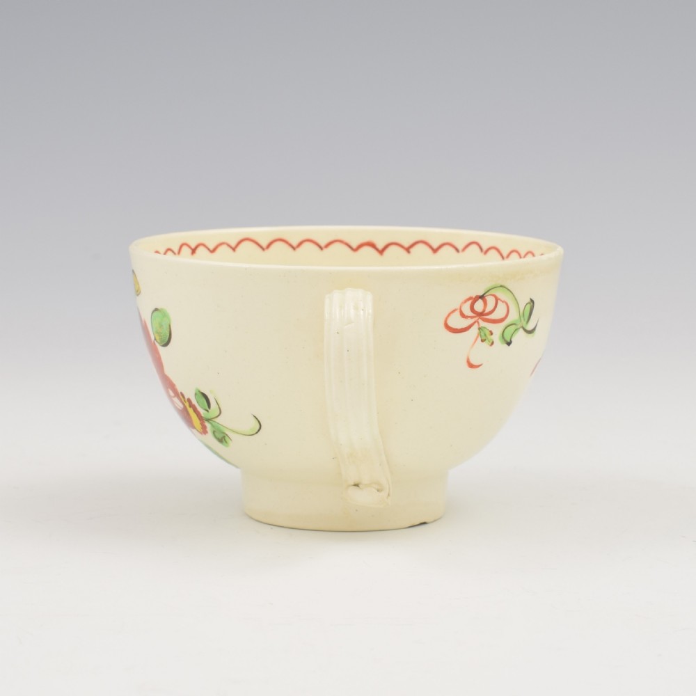 An antique creamware tea cup and saucer featuring hand-painted red, green, and yellow floral motifs in the Kings Rose pattern against a light cream backdrop. View VI