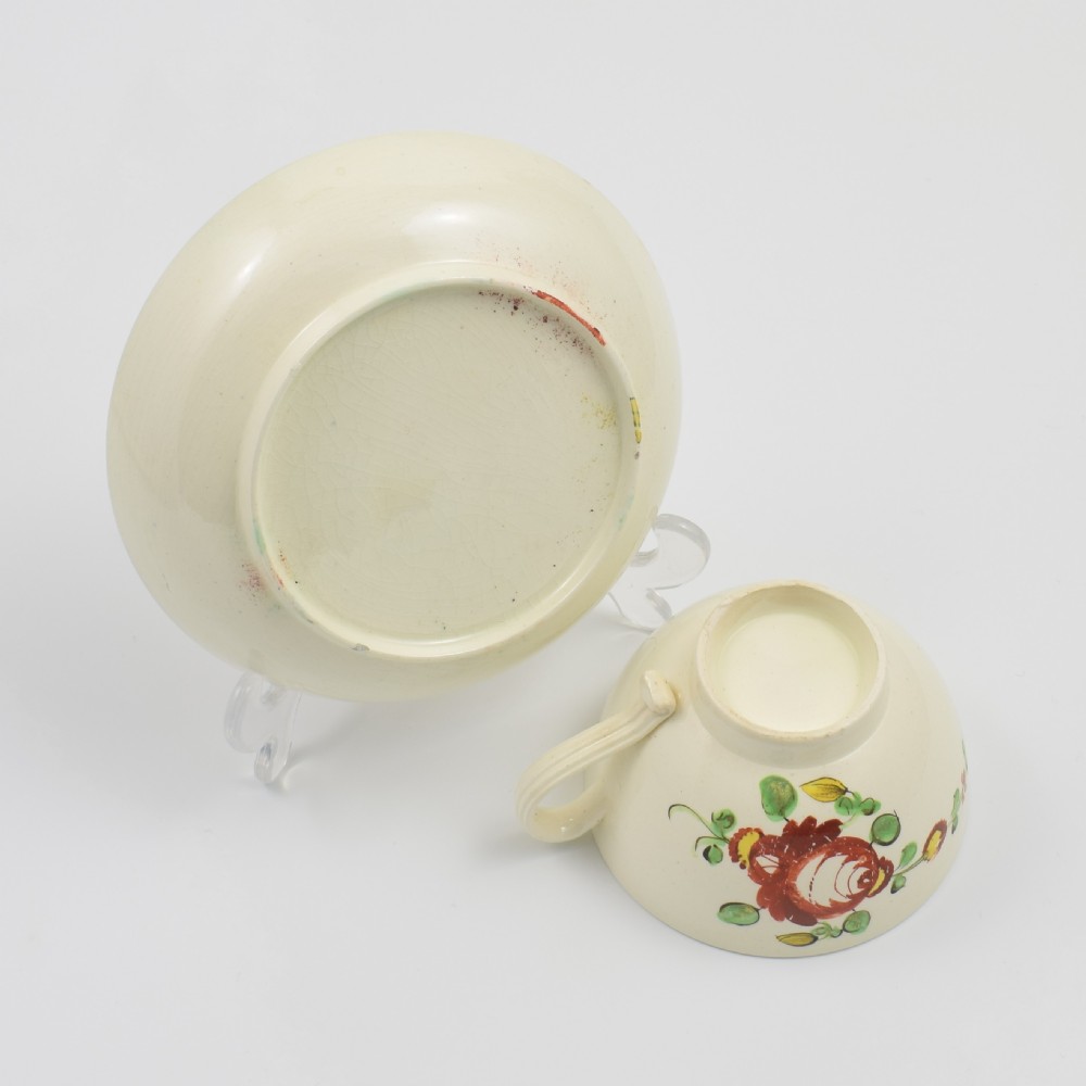 An antique creamware tea cup and saucer featuring hand-painted red, green, and yellow floral motifs in the Kings Rose pattern against a light cream backdrop. View VII