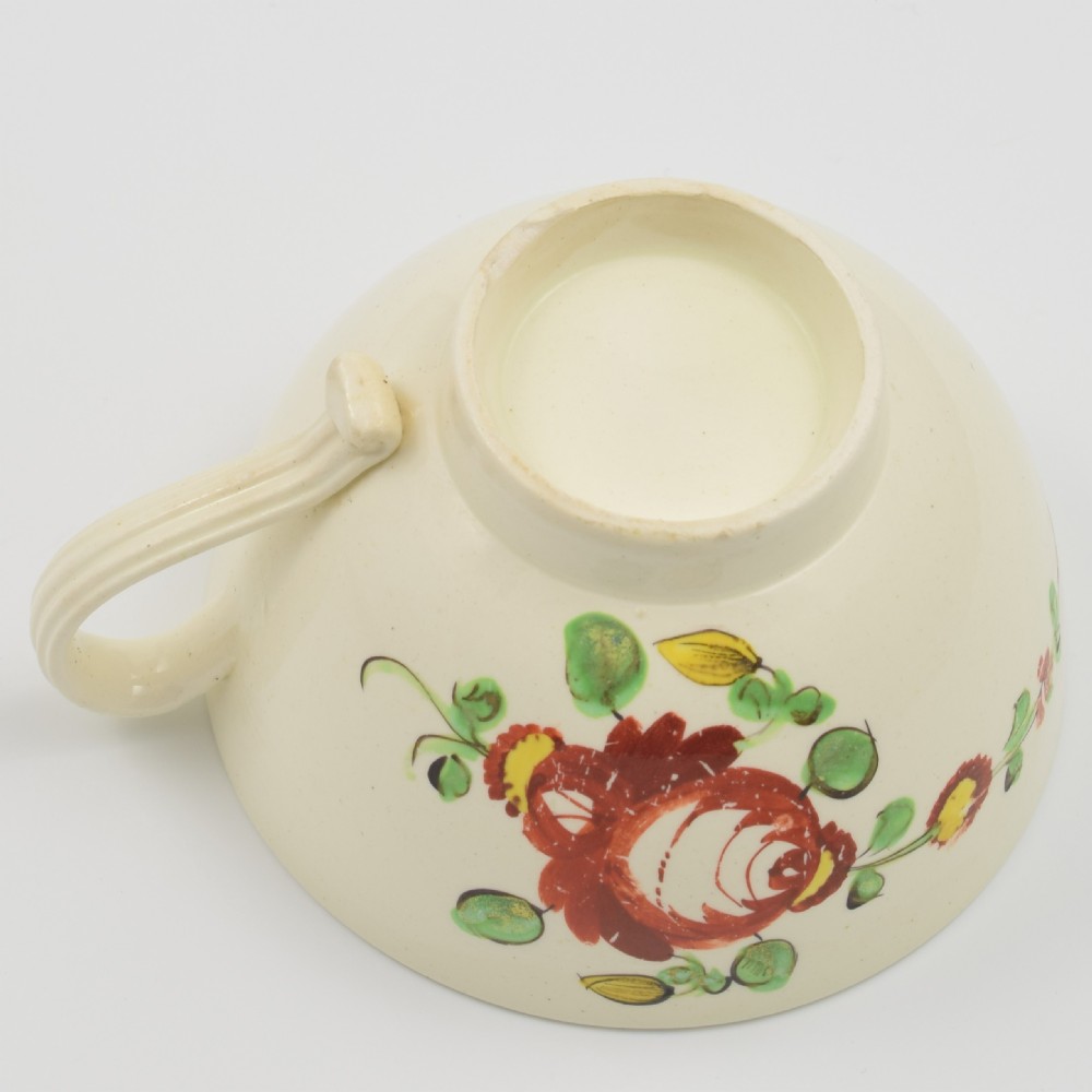 An antique creamware tea cup and saucer featuring hand-painted red, green, and yellow floral motifs in the Kings Rose pattern against a light cream backdrop. View VIII