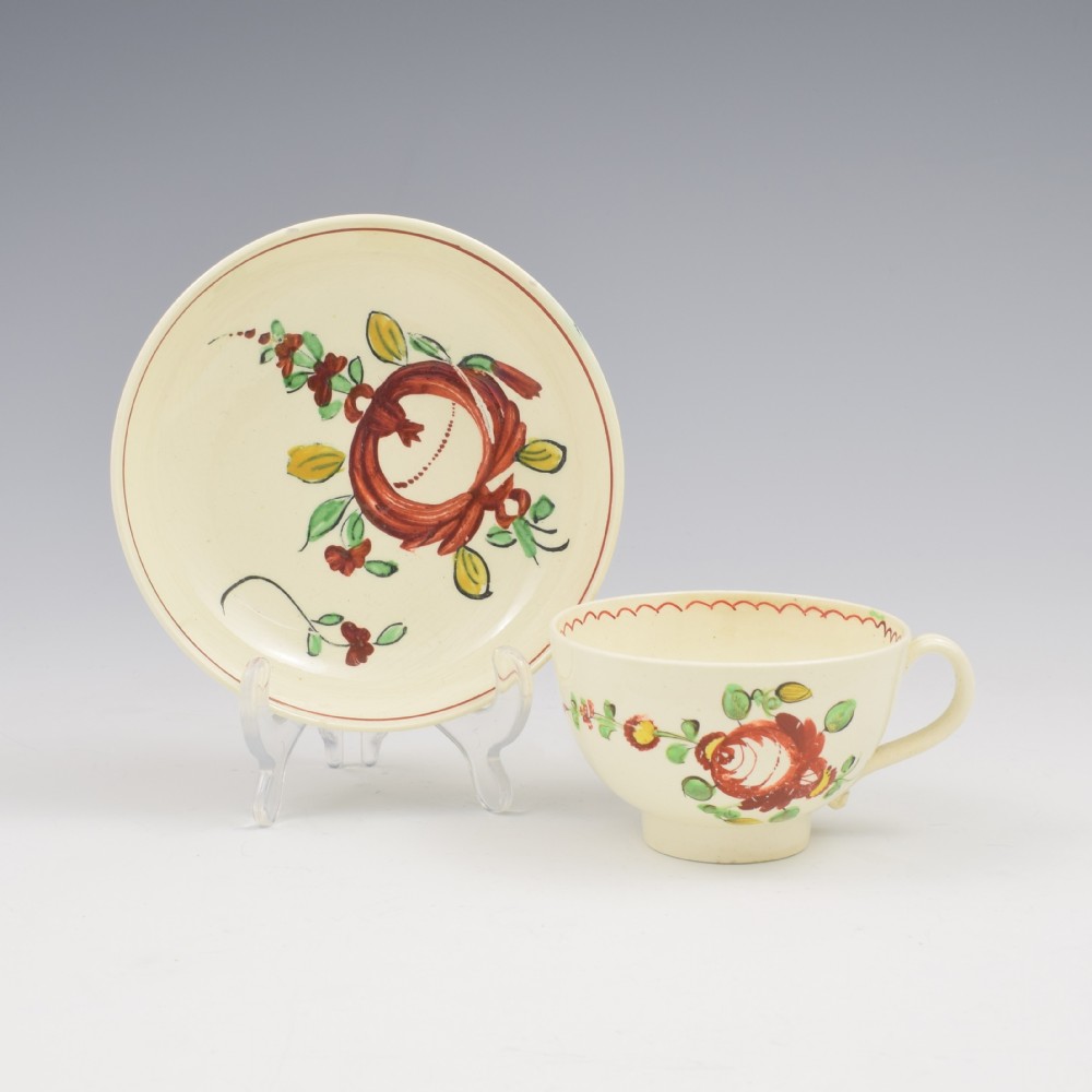 An antique creamware tea cup and saucer featuring hand-painted red, green, and yellow floral motifs in the Kings Rose pattern against a light cream backdrop.