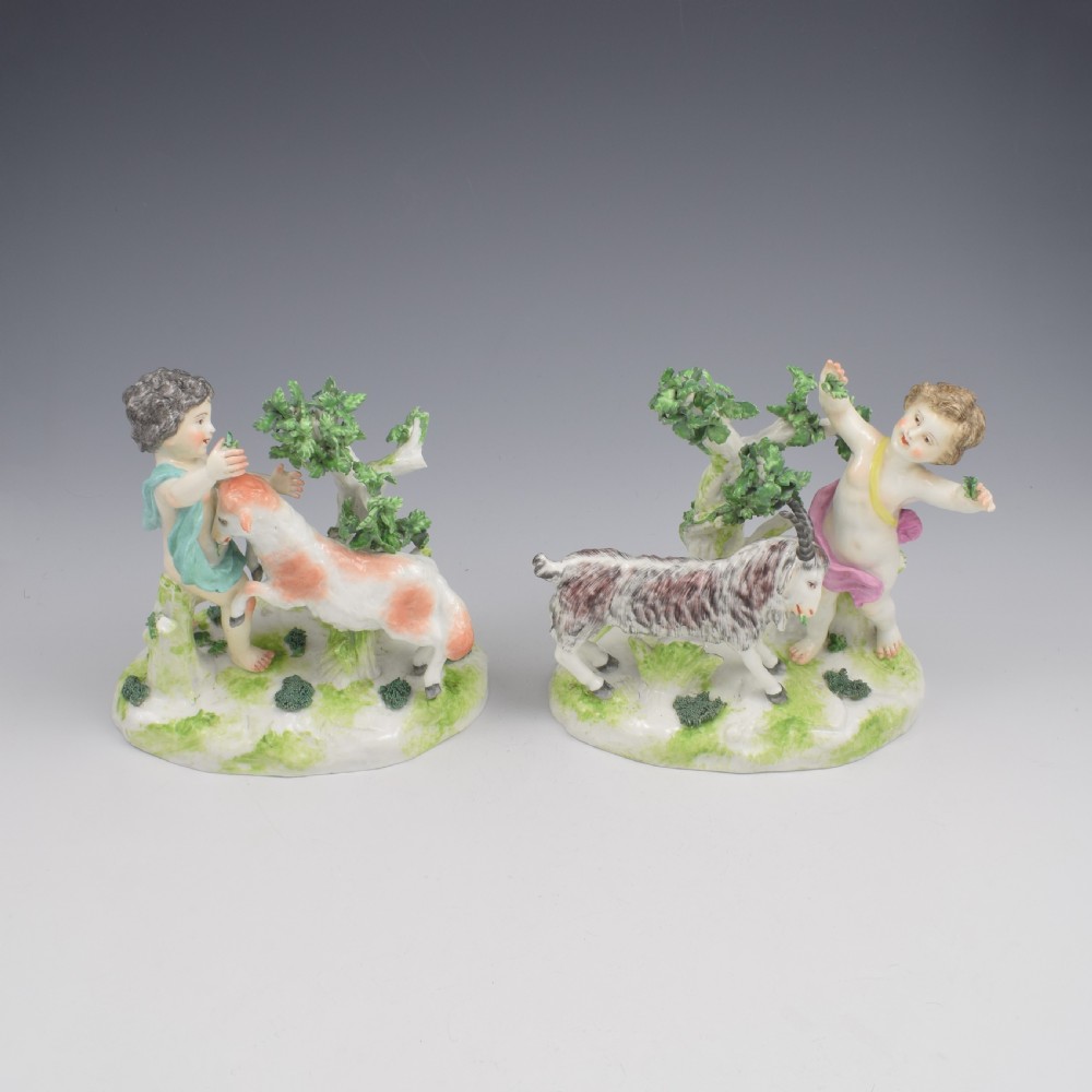 A pair of hand-painted porcelain figures featuring young putti standing alongside goats amidst green leafy foliage on rounded naturalistic bases.