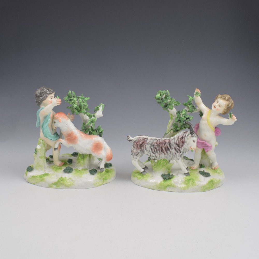A pair of hand-painted porcelain figures featuring young putti standing alongside goats amidst green leafy foliage on rounded naturalistic bases. View II