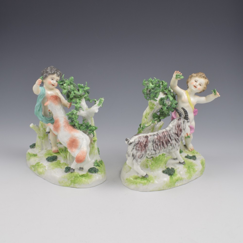 A pair of hand-painted porcelain figures featuring young putti standing alongside goats amidst green leafy foliage on rounded naturalistic bases. View III