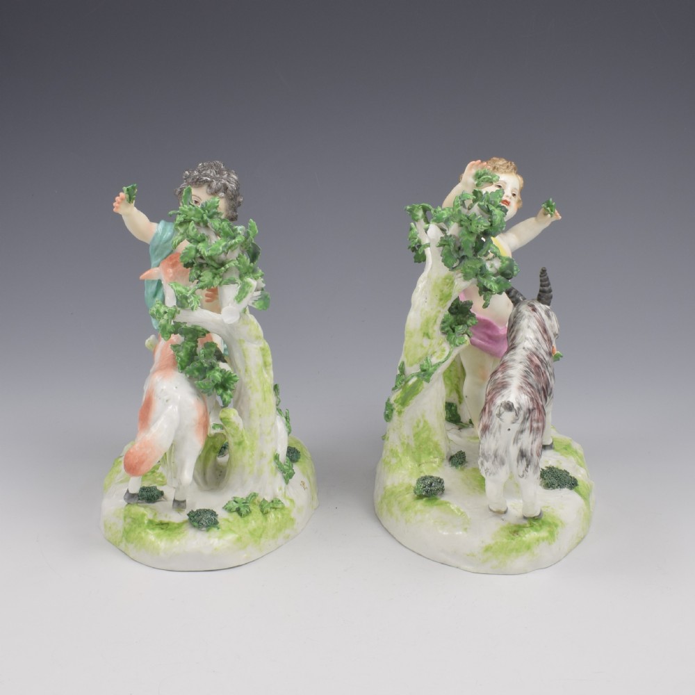 A pair of hand-painted porcelain figures featuring young putti standing alongside goats amidst green leafy foliage on rounded naturalistic bases. View IV