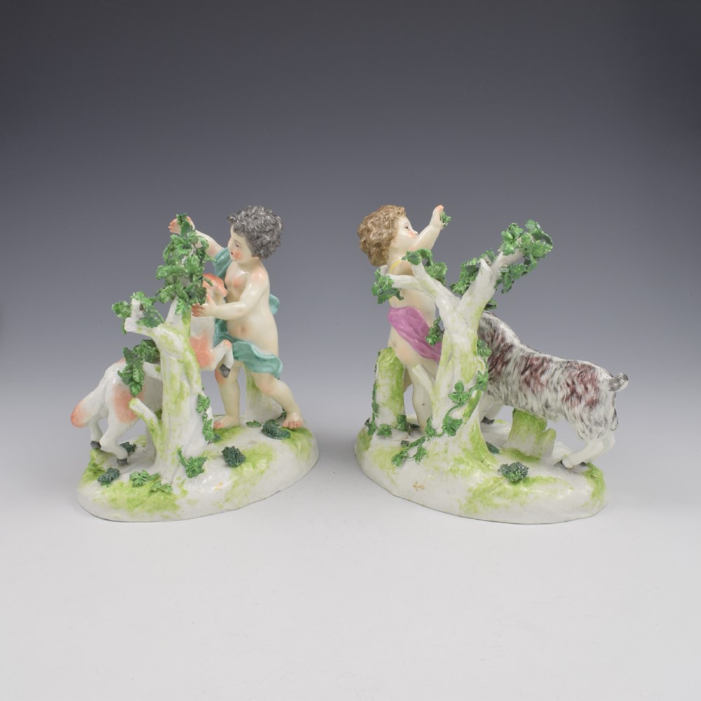 A pair of hand-painted porcelain figures featuring young putti standing alongside goats amidst green leafy foliage on rounded naturalistic bases. View V