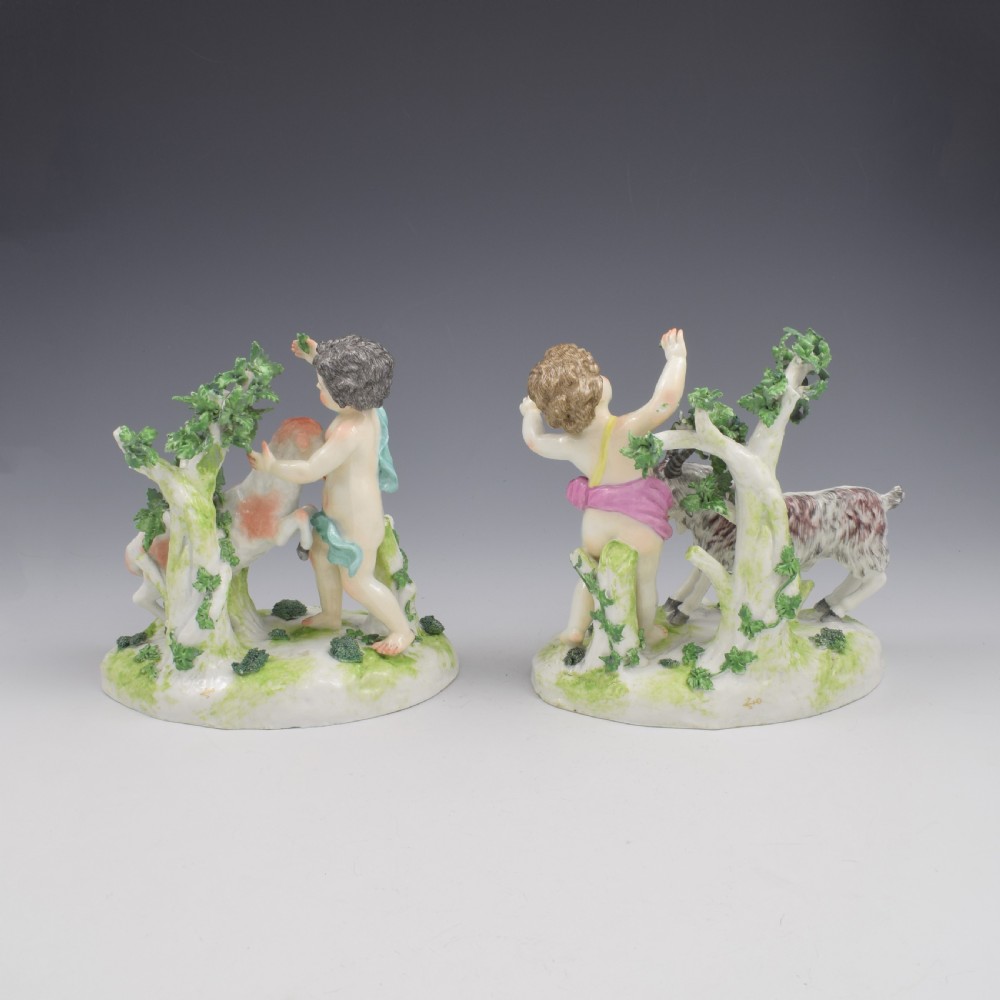 A pair of hand-painted porcelain figures featuring young putti standing alongside goats amidst green leafy foliage on rounded naturalistic bases. View VI