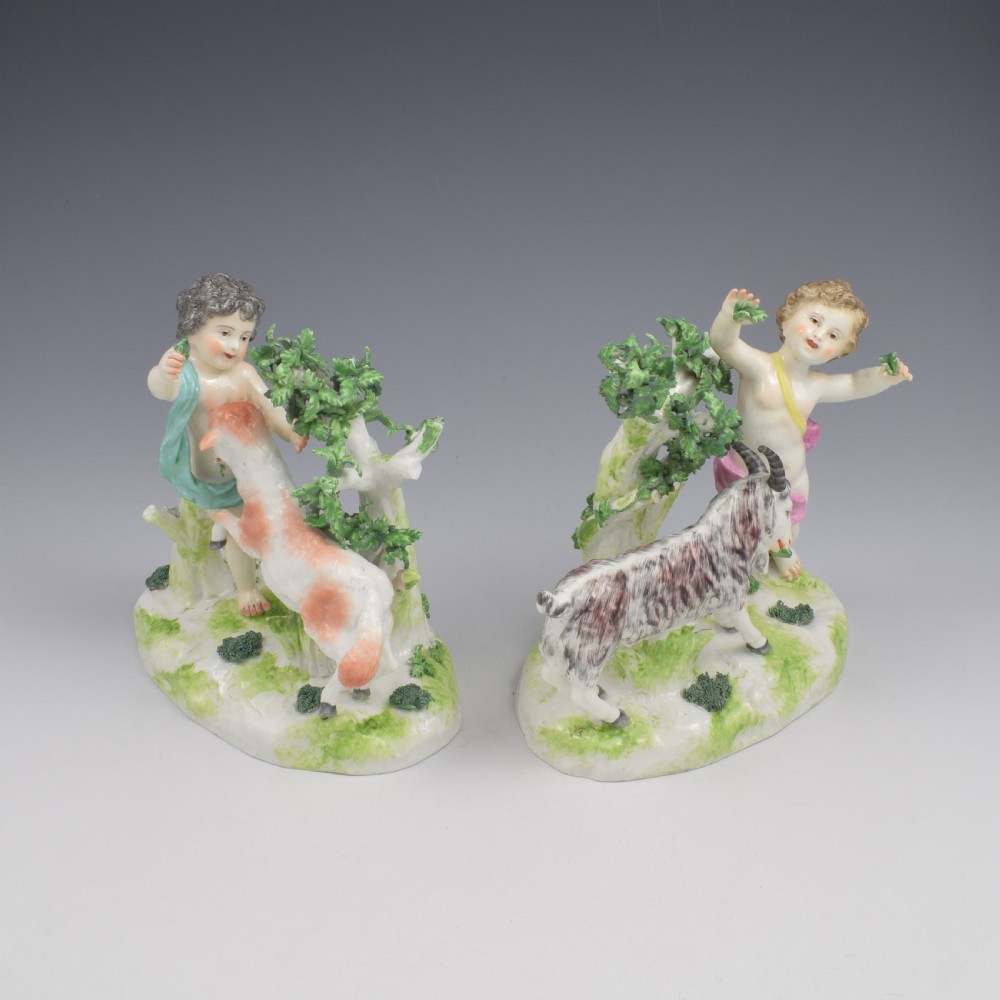 A pair of hand-painted porcelain figures featuring young putti standing alongside goats amidst green leafy foliage on rounded naturalistic bases. View VII
