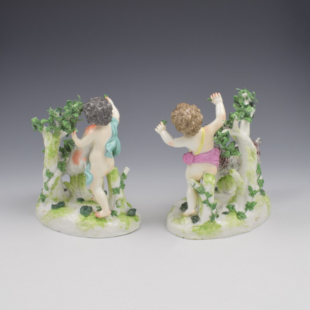 A pair of hand-painted porcelain figures featuring young putti standing alongside goats amidst green leafy foliage on rounded naturalistic bases. View VIII