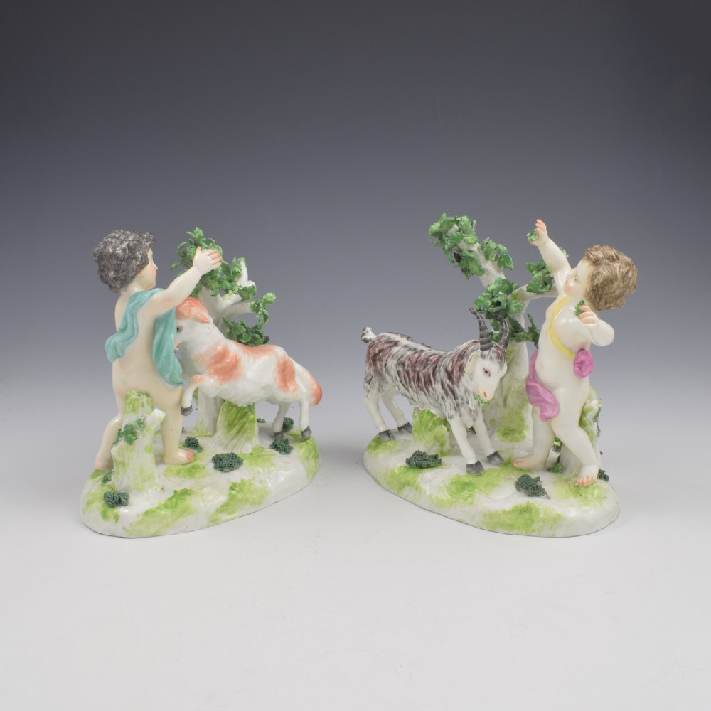 A pair of hand-painted porcelain figures featuring young putti standing alongside goats amidst green leafy foliage on rounded naturalistic bases. View IX