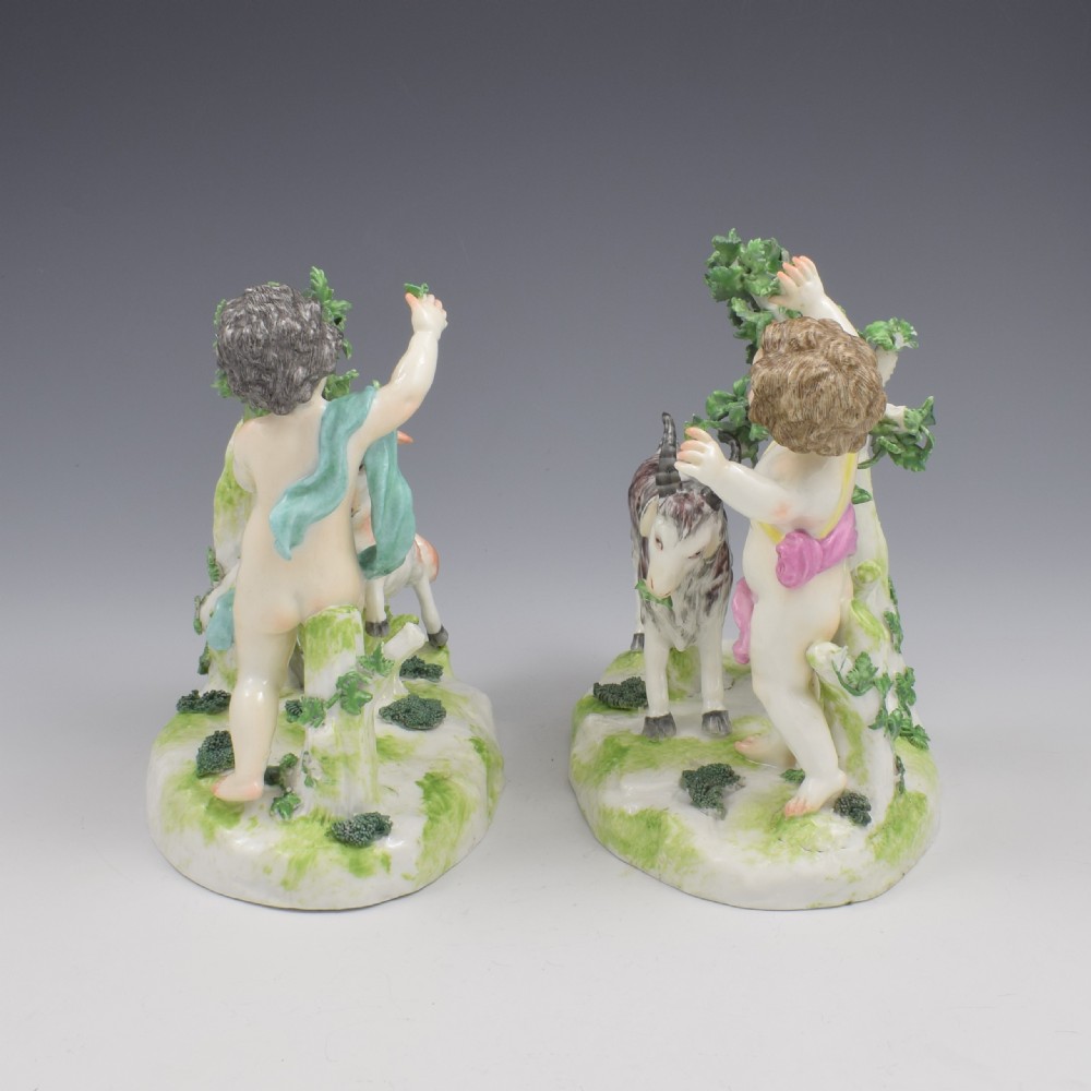 A pair of hand-painted porcelain figures featuring young putti standing alongside goats amidst green leafy foliage on rounded naturalistic bases. View X