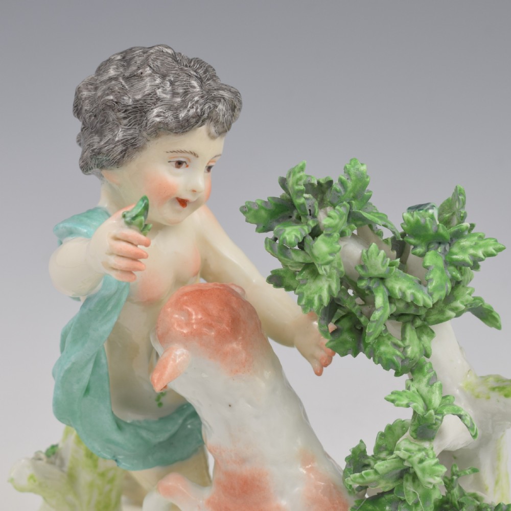 A pair of hand-painted porcelain figures featuring young putti standing alongside goats amidst green leafy foliage on rounded naturalistic bases. View XI