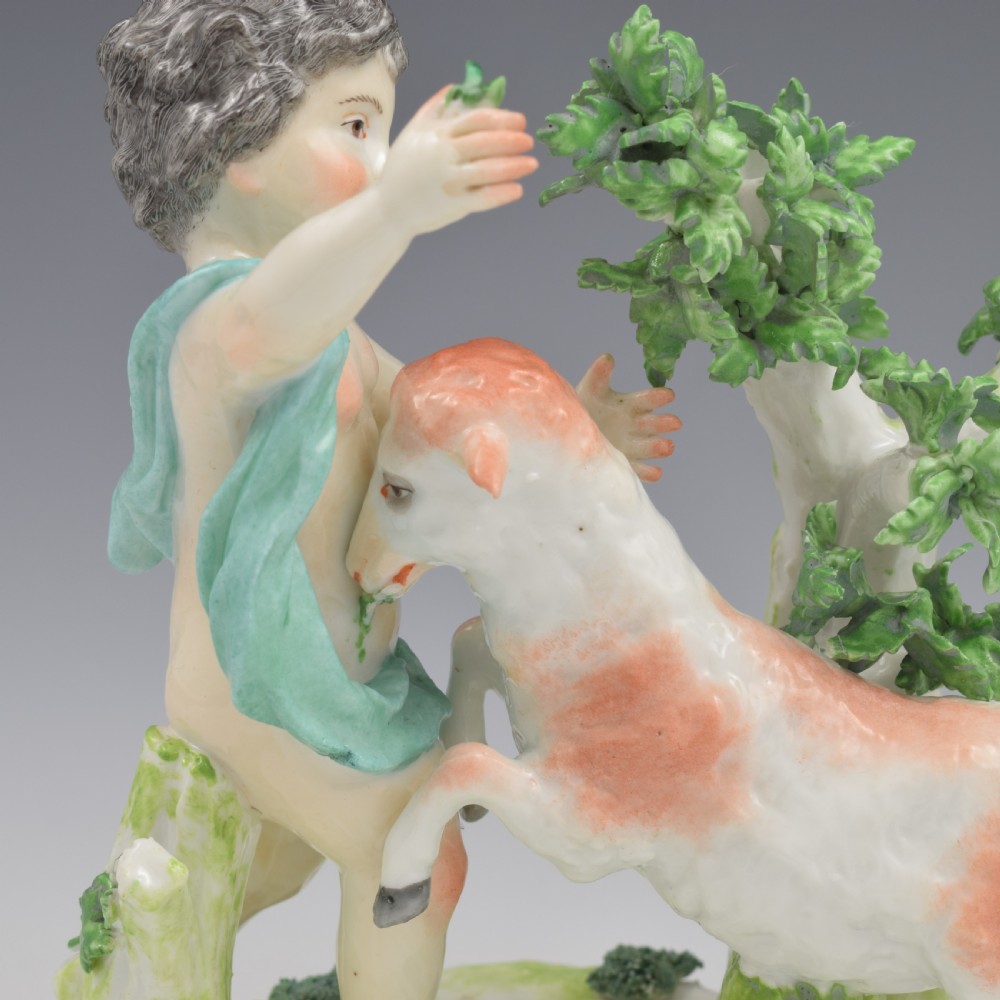 A pair of hand-painted porcelain figures featuring young putti standing alongside goats amidst green leafy foliage on rounded naturalistic bases. View XII
