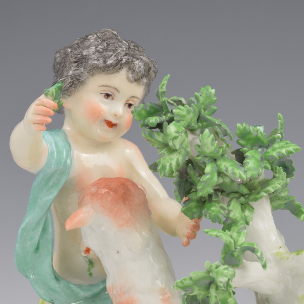 A pair of hand-painted porcelain figures featuring young putti standing alongside goats amidst green leafy foliage on rounded naturalistic bases. View XIII