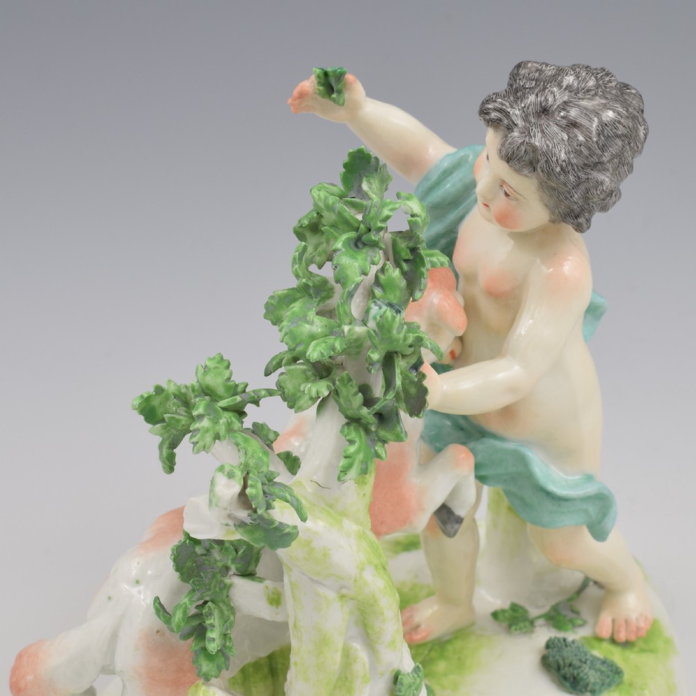 A pair of hand-painted porcelain figures featuring young putti standing alongside goats amidst green leafy foliage on rounded naturalistic bases. View XIV