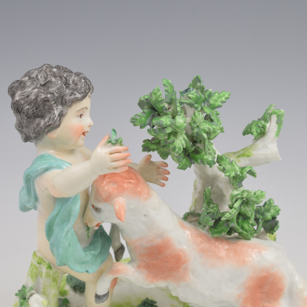 A pair of hand-painted porcelain figures featuring young putti standing alongside goats amidst green leafy foliage on rounded naturalistic bases. View XV