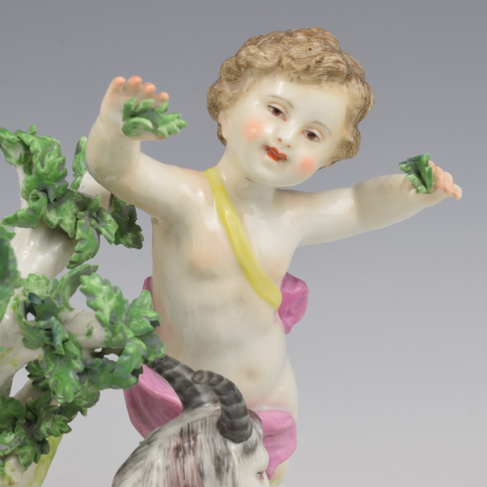 A pair of hand-painted porcelain figures featuring young putti standing alongside goats amidst green leafy foliage on rounded naturalistic bases. View XVII