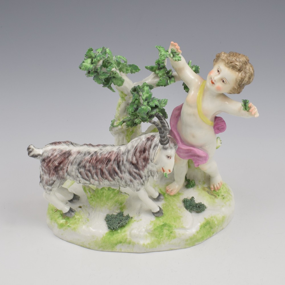 A pair of hand-painted porcelain figures featuring young putti standing alongside goats amidst green leafy foliage on rounded naturalistic bases. View XVIII