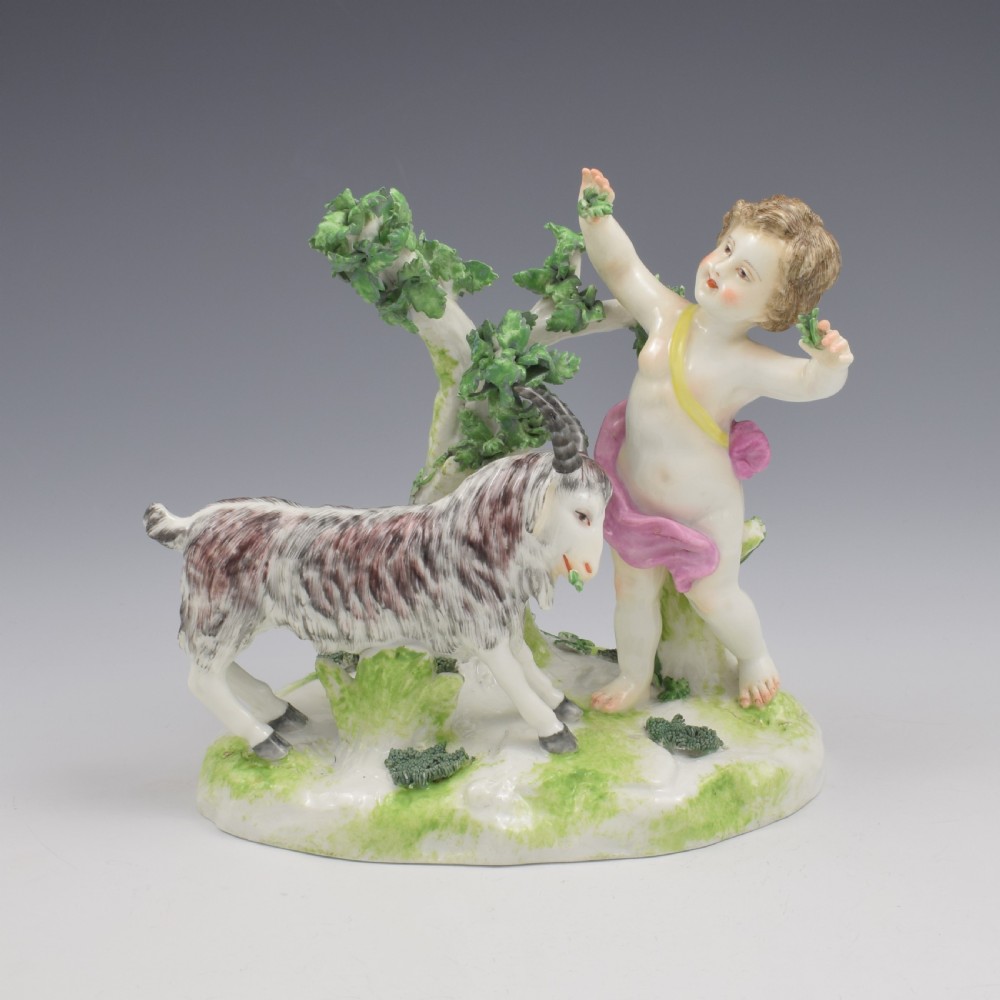 A pair of hand-painted porcelain figures featuring young putti standing alongside goats amidst green leafy foliage on rounded naturalistic bases. View XXI