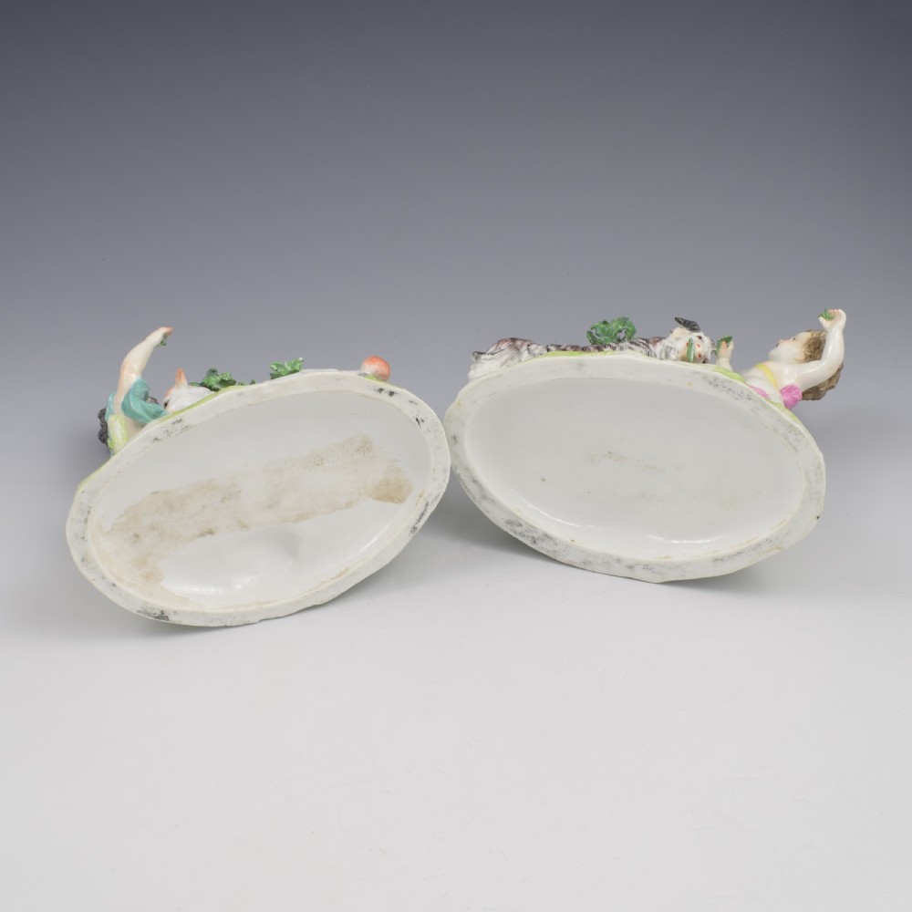 A pair of hand-painted porcelain figures featuring young putti standing alongside goats amidst green leafy foliage on rounded naturalistic bases. View XXIII