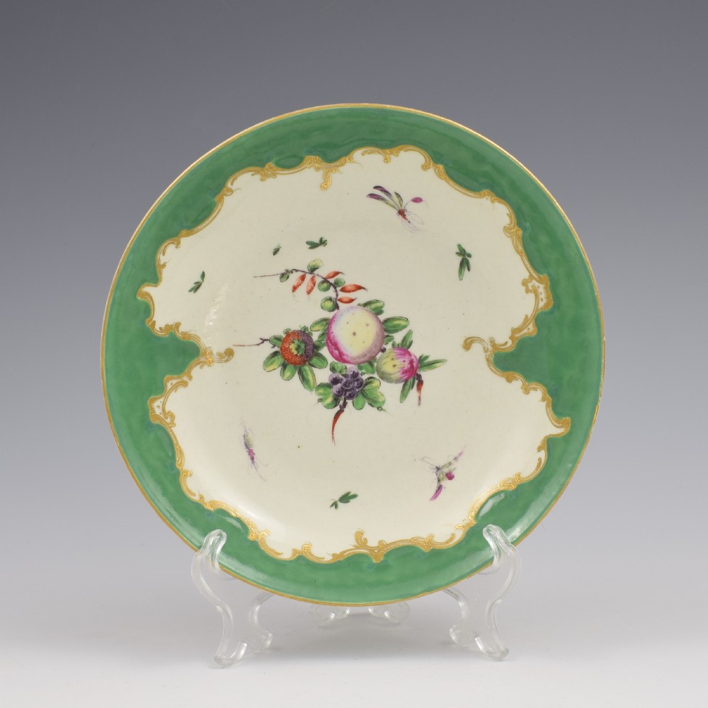 A circular eighteenth-century porcelain dish featuring an apple green border with gilded rococo scrolling surrounding a white centre painted with fruit and insects.