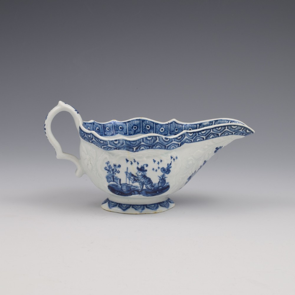 An eighteenth-century English soft-paste porcelain sauce boat decorated with underglaze cobalt blue Chinoiserie figures, a scalloped rim, and a molded handle.