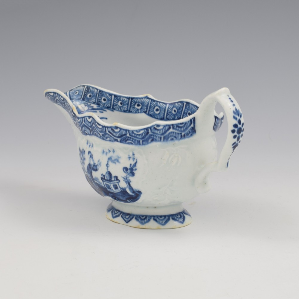 An eighteenth-century English soft-paste porcelain sauce boat decorated with underglaze cobalt blue Chinoiserie figures, a scalloped rim, and a molded handle. View V