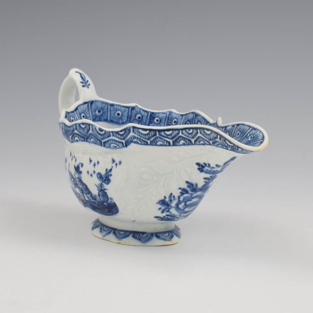 An eighteenth-century English soft-paste porcelain sauce boat decorated with underglaze cobalt blue Chinoiserie figures, a scalloped rim, and a molded handle. View X