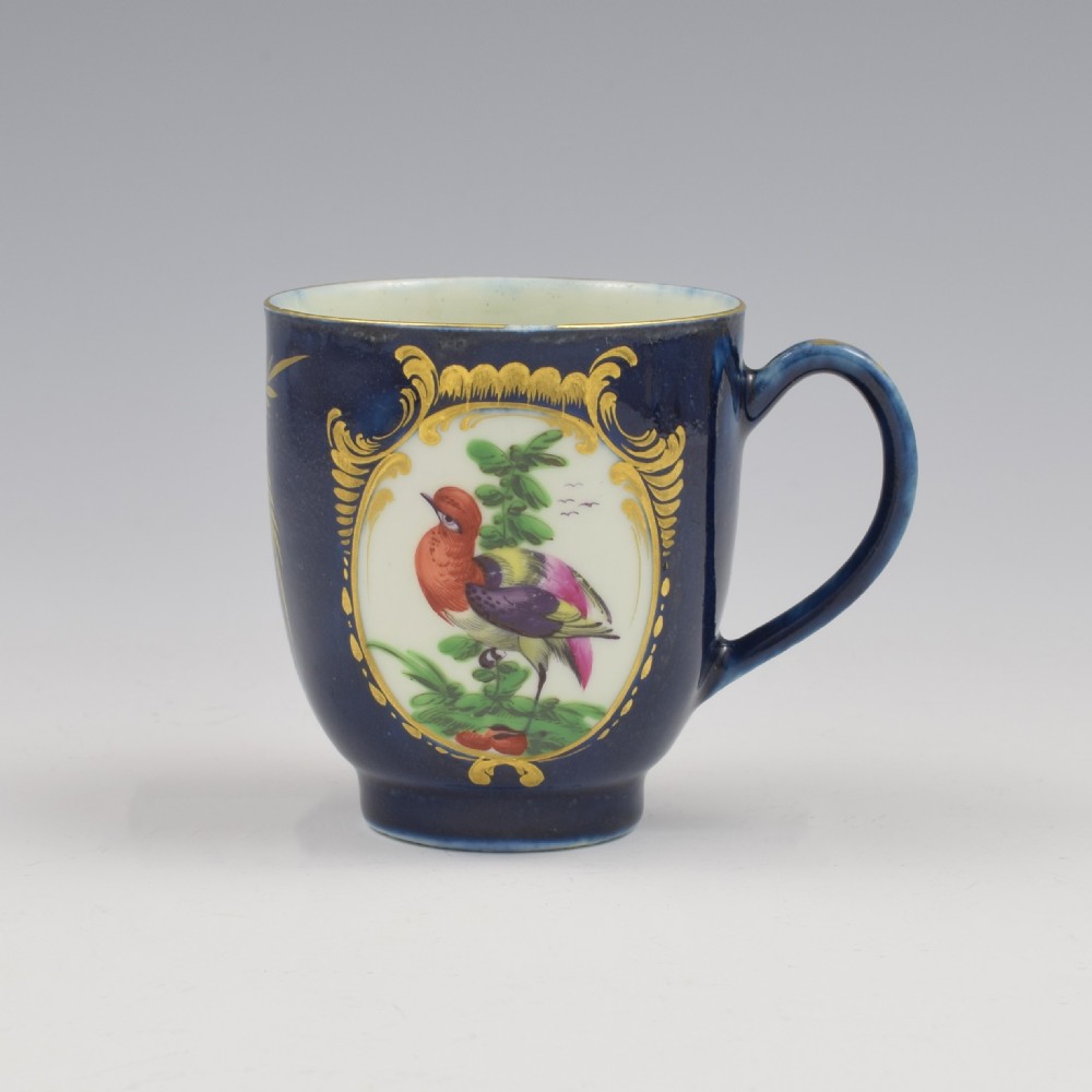 A dark blue soft-paste porcelain coffee cup decorated with a gilded cartouche enclosing a painted exotic bird on a foliage branch.