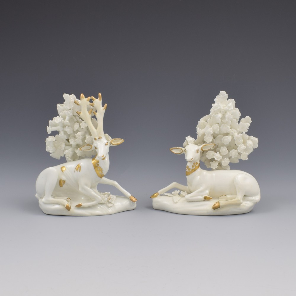A pair of white glazed porcelain figures representing a recumbent stag and doe with gilded accents set against floral bocage tree backgrounds.