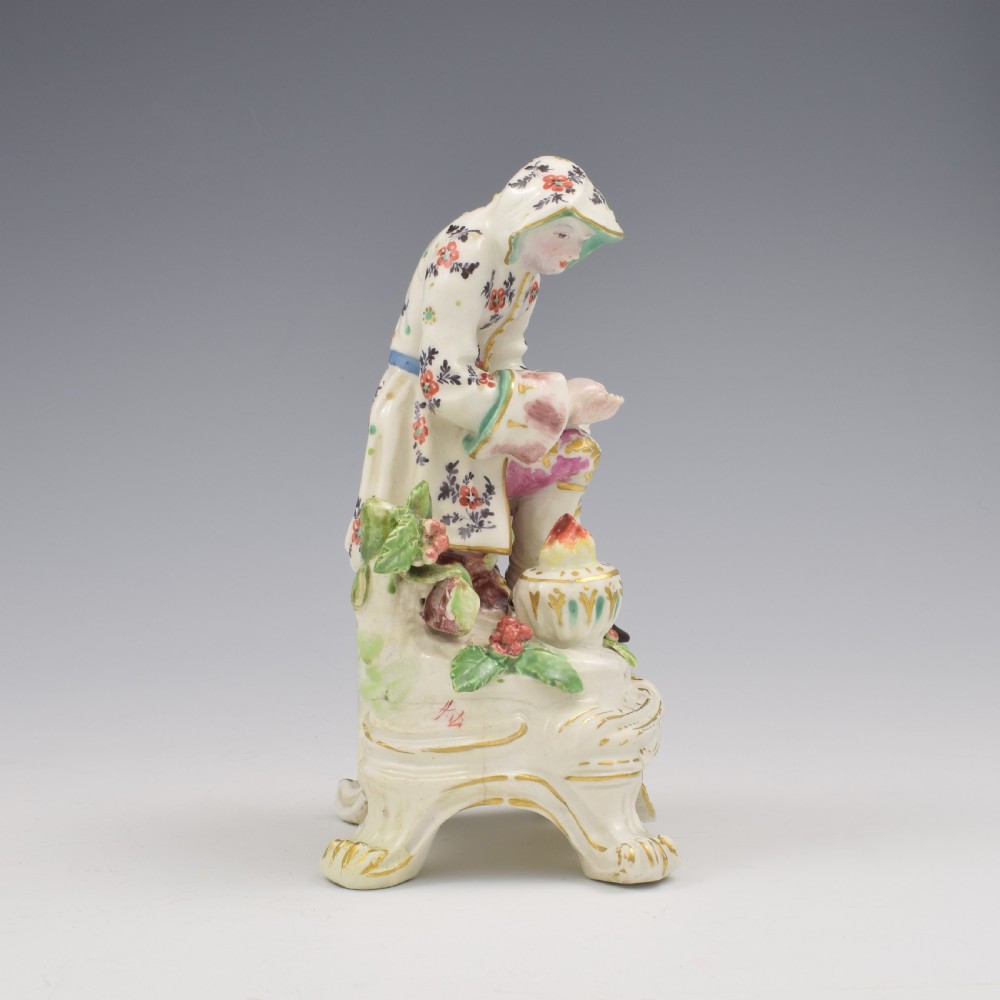 An 18th-century Bow porcelain figure representing Winter, featuring a hooded figure seated on a gilt scrollwork base warming hands over a brazier. View III