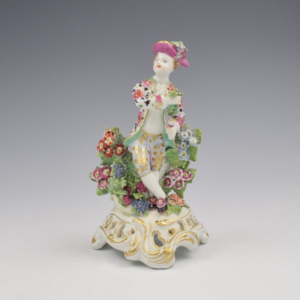 An 18th-century Bow soft-paste porcelain figure depicting an allegorical youth standing beside floral bocage on a pierced gilded scroll base.