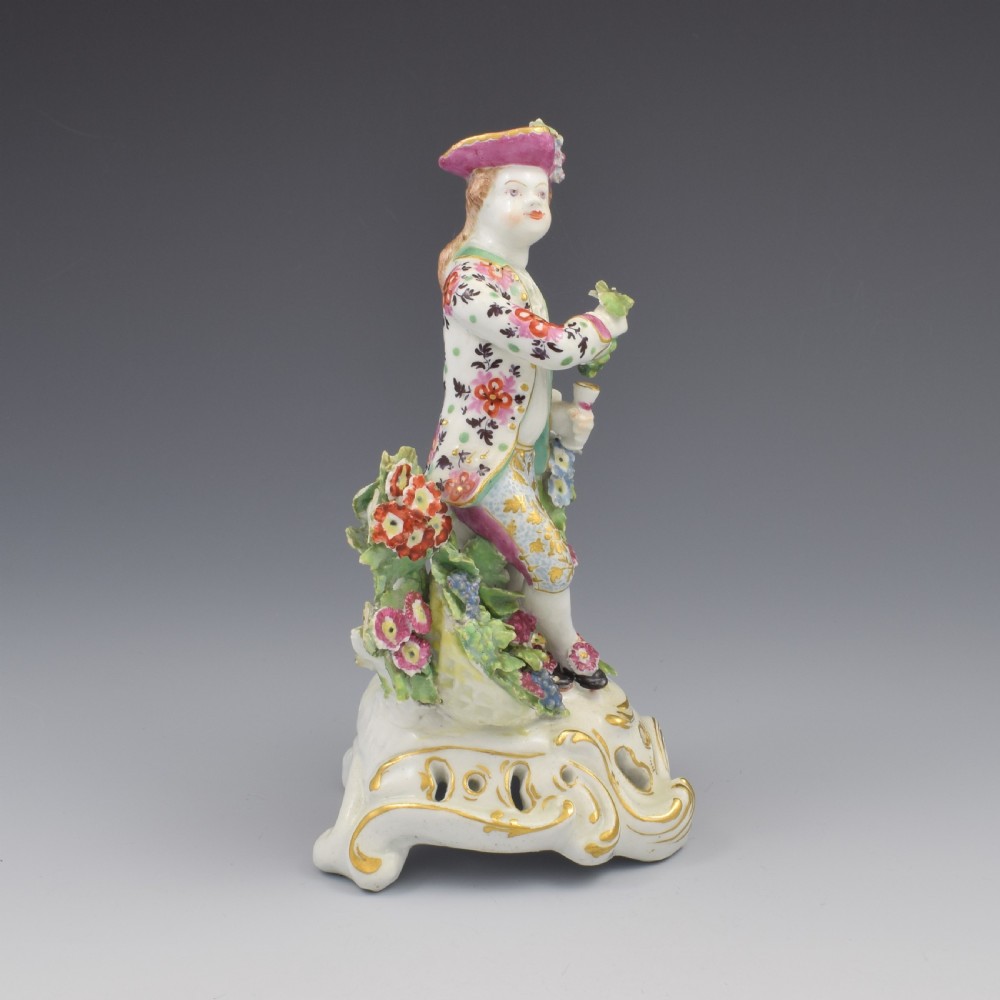 An 18th-century Bow soft-paste porcelain figure depicting an allegorical youth standing beside floral bocage on a pierced gilded scroll base. View II