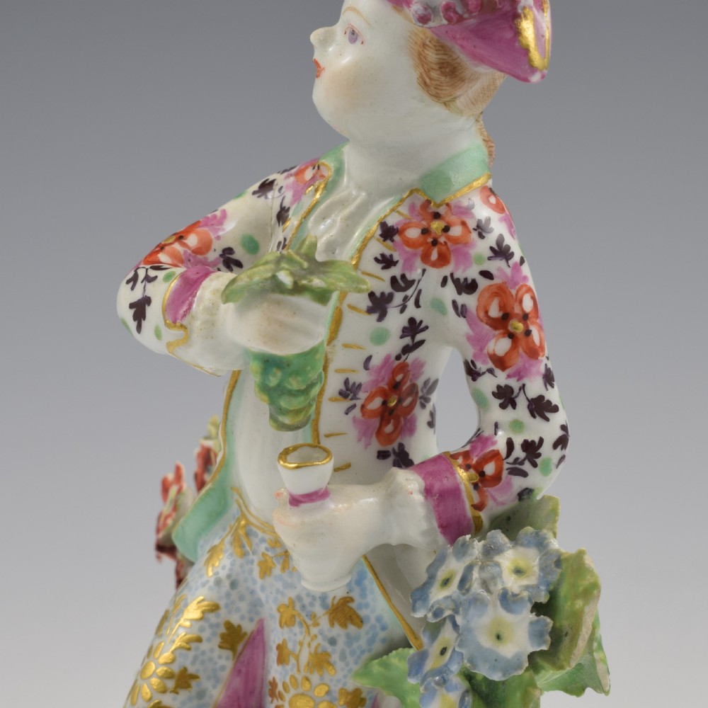 An 18th-century Bow soft-paste porcelain figure depicting an allegorical youth standing beside floral bocage on a pierced gilded scroll base. View X