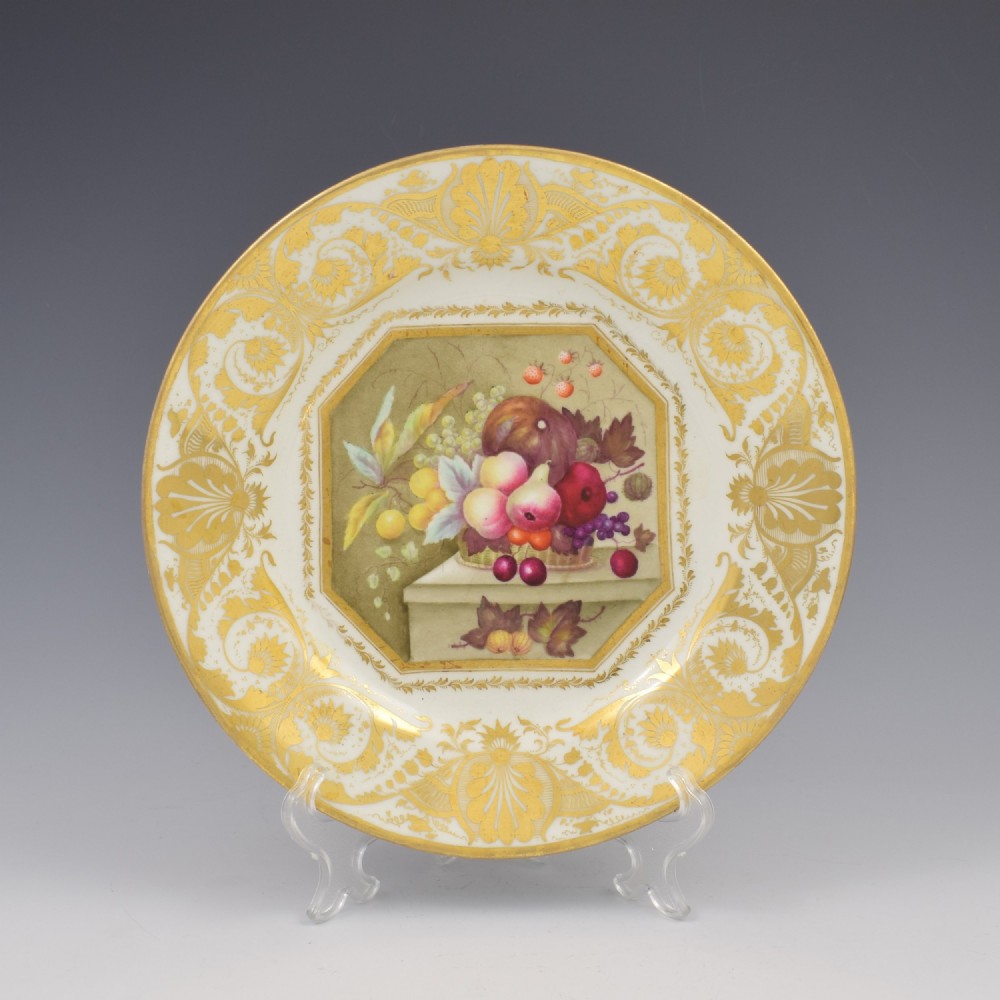 A Regency era Derby porcelain dessert plate featuring a central octagonal panel hand-painted with still-life fruit on a stone ledge, bordered by intricate gold gilding.