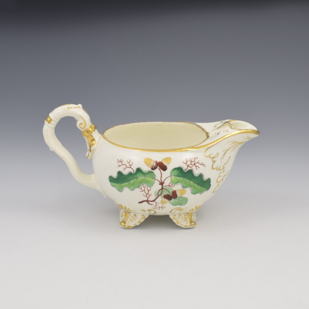 A cream-coloured Regency porcelain jug featuring hand-painted green oak leaves, brown acorns, moulded feet, and gilded trim.