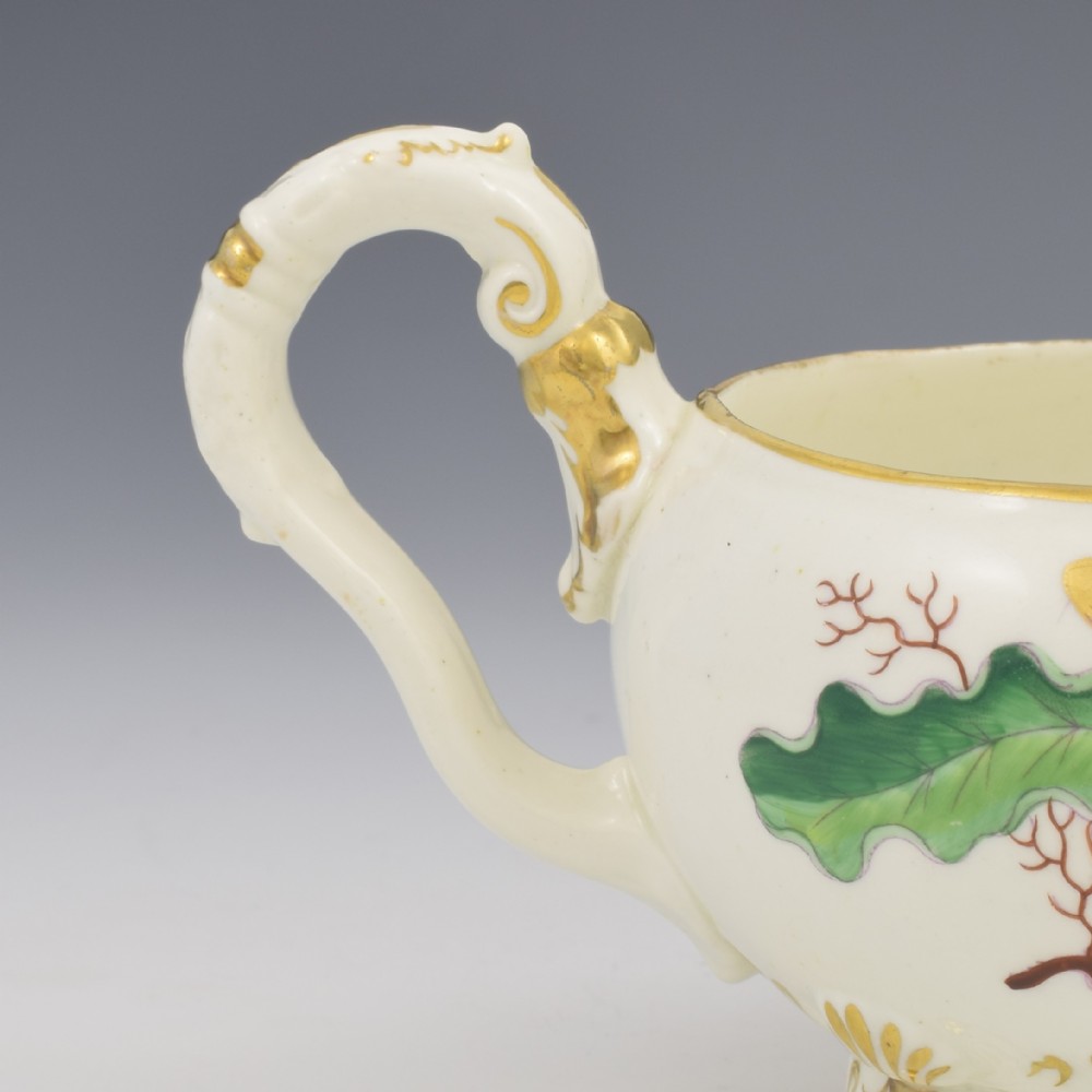 A cream-coloured Regency porcelain jug featuring hand-painted green oak leaves, brown acorns, moulded feet, and gilded trim. View X